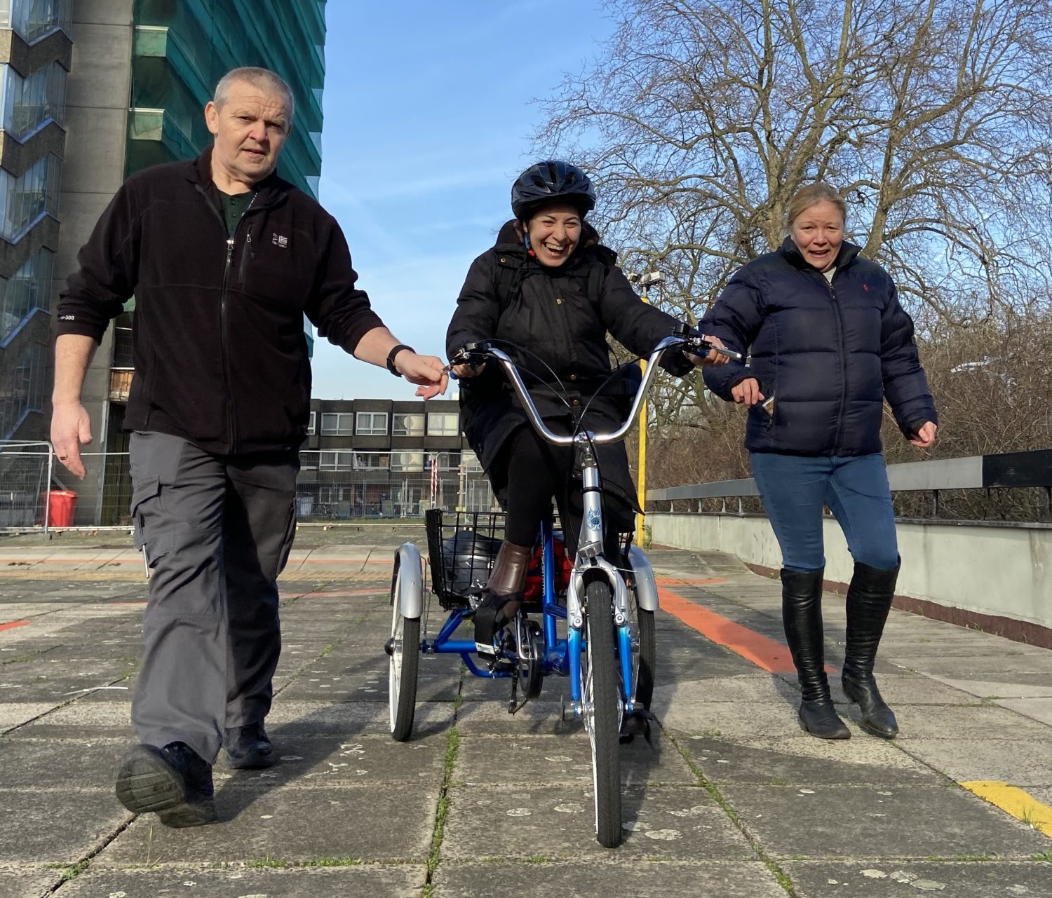 Two staff support a laughing Learning Disabilities client, by walking either side of her and enabling her to take an enjoyable spin on her trike in safety